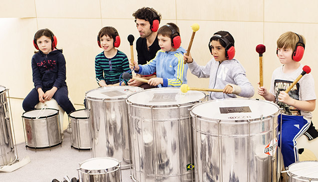 Un intervenant assis derrière 5 écoliers, dont 3 jouent de la cuíca, percussion brésilienne