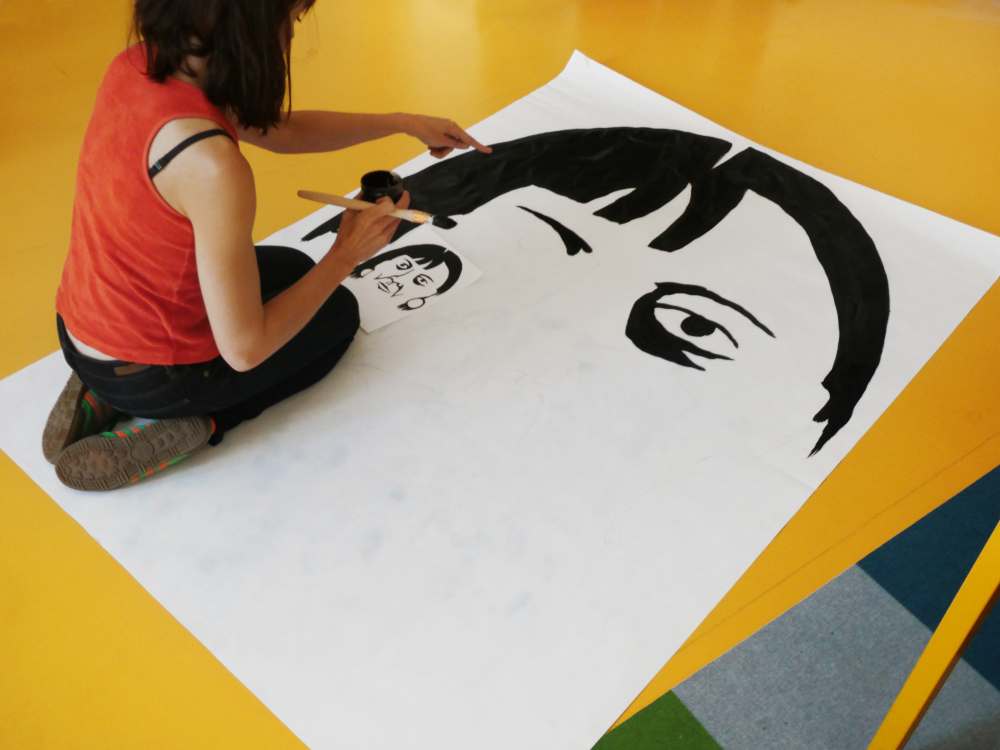 À genoux sur une grande feuille A0, une femme de dos copie au pinceau noir un portrait au format A5