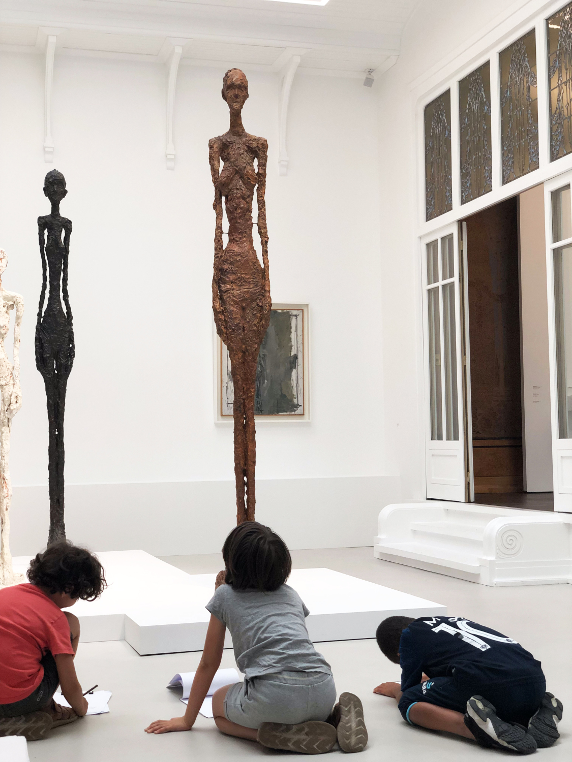 3 scolaires dessinent à genoux devant des sculptures de Giacometti