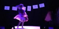 Sous une lumière bleue, une comédienne en mouvement sur scène devant une guirlande de dessins